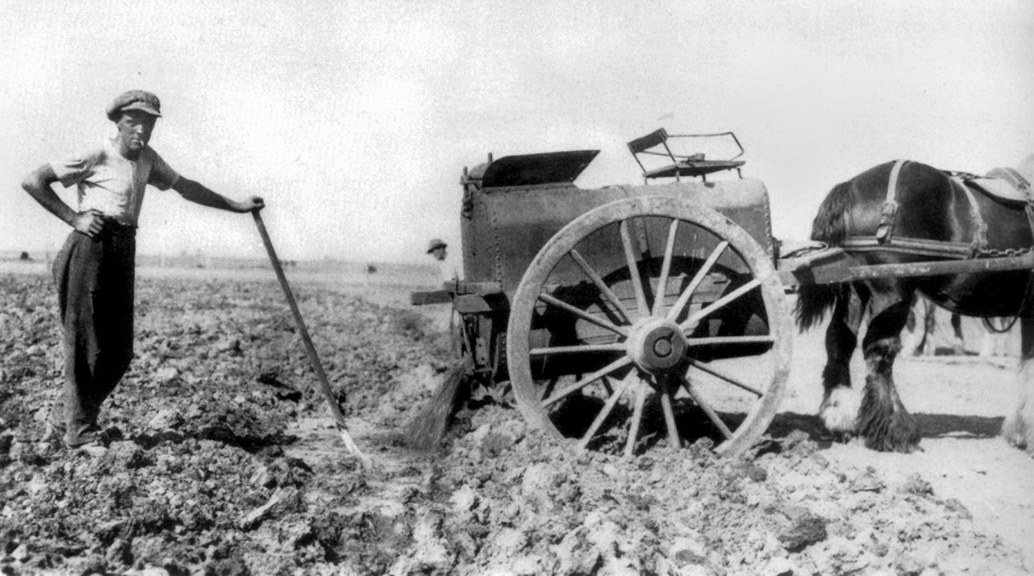 a black and white photo of a man with a shovel standing in a field next to a horse and cart