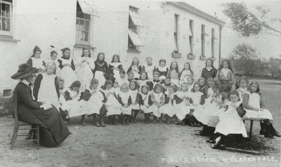 Many uniformed young girls taking an outdoors sewing class, overseen by a teacher