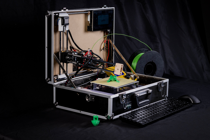 A photo of an open black suitcase containing 3D printer equipment and wiring, and two small green 3D printed models. A black keyboard and a black mouse sit in front of the suitcase.