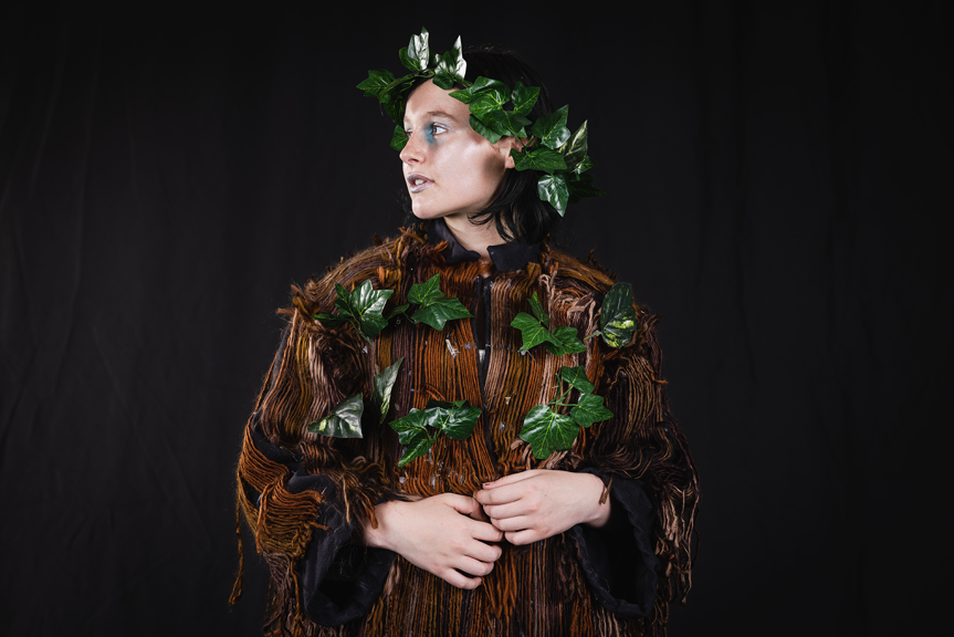 A person wearing a garment covered in brown thread and leaves, with a halo of leaves on their head. They are looking to the left of the frame.