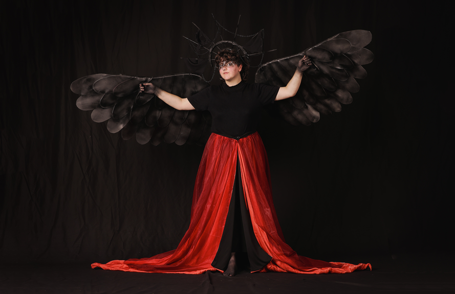 A person wears a long red skirt which is draped on the floor, with large black wings and a halo attached to their arms and head.