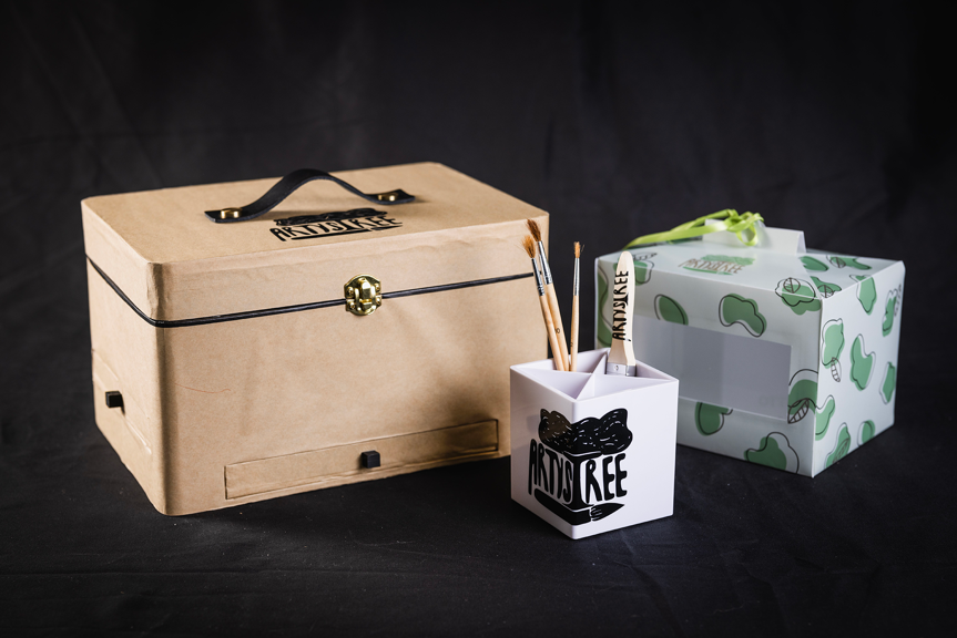 Three art supply storage units, which all have logos reading ‘Artystree’. There is a large card box with a handle and gold hinge,  paintbrush holder, and green and white packaging.