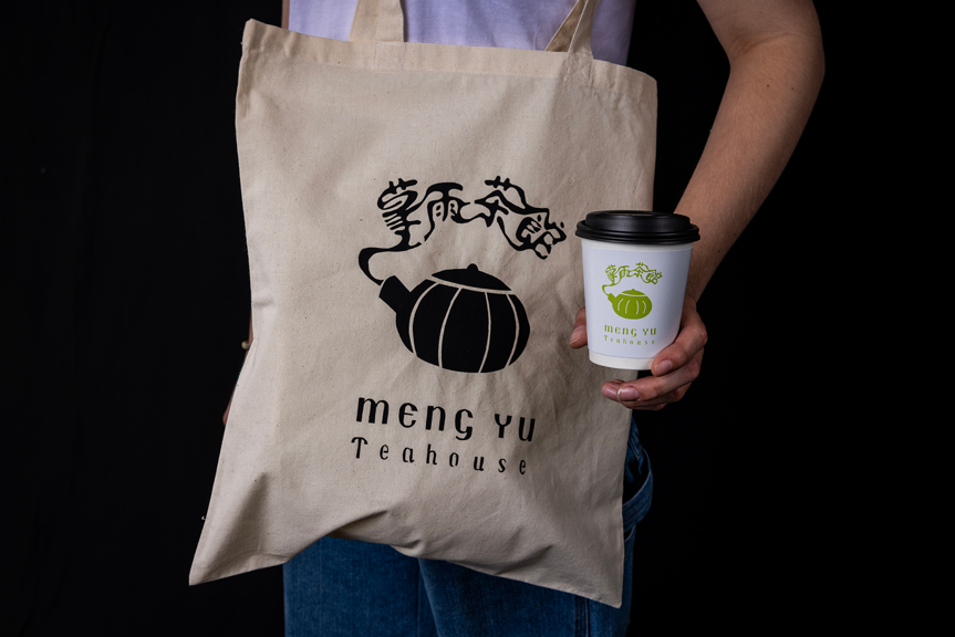A person with a branded Meng Yu Teahouse tote bag over their shoulder. They hold a takeaway cup with a green Meng Yu Teahouse logo on it.
