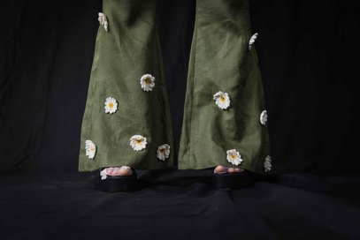 A close up photograph of the final product, showing the bottom of wide trouser legs. The trousers are olive green and have knitted daisies attached.
