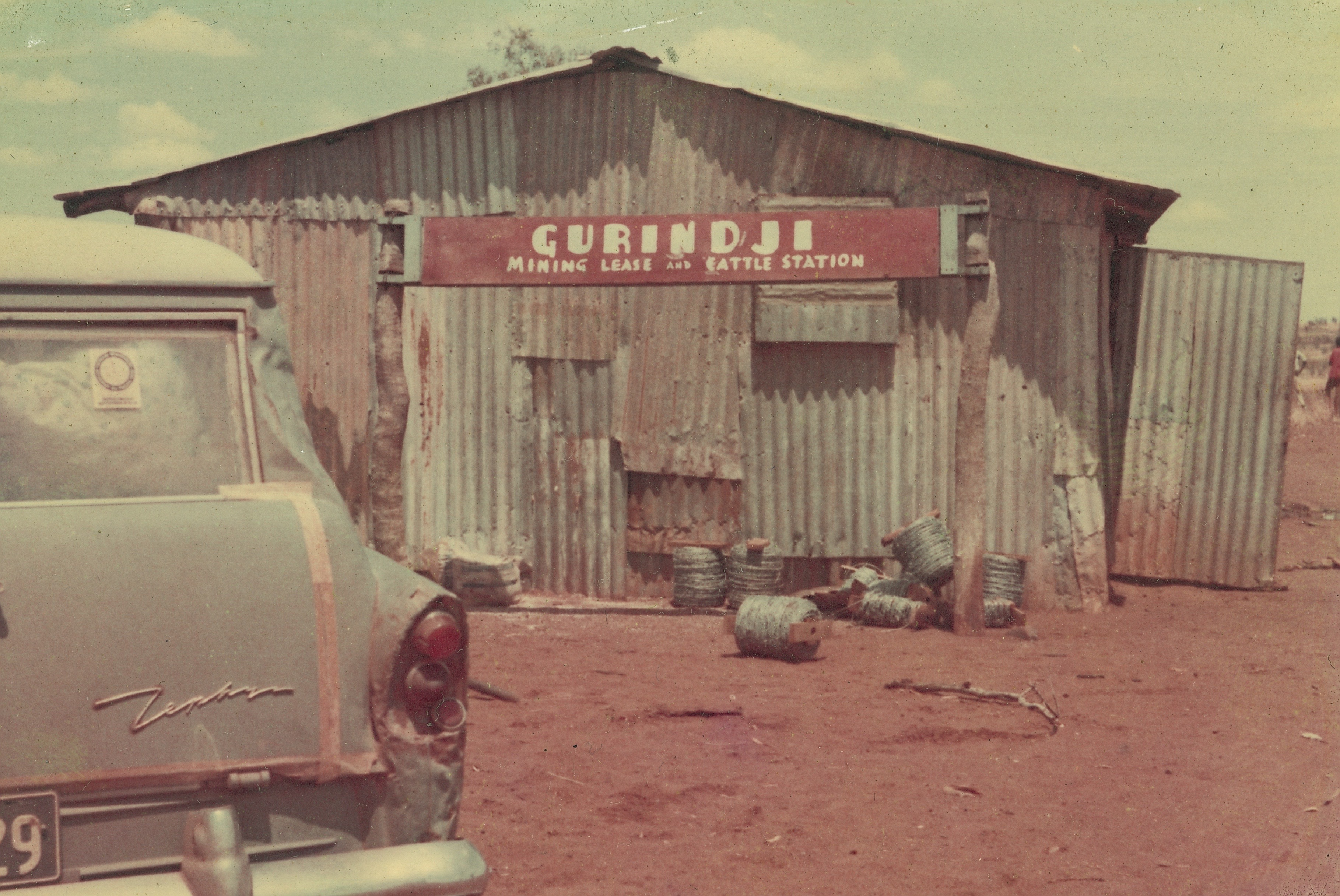 Rusty iron shed bearing a sign reading 'Gurindji Mining Lease and Cattle Station'. A Ford Zephyr is parked out the front.