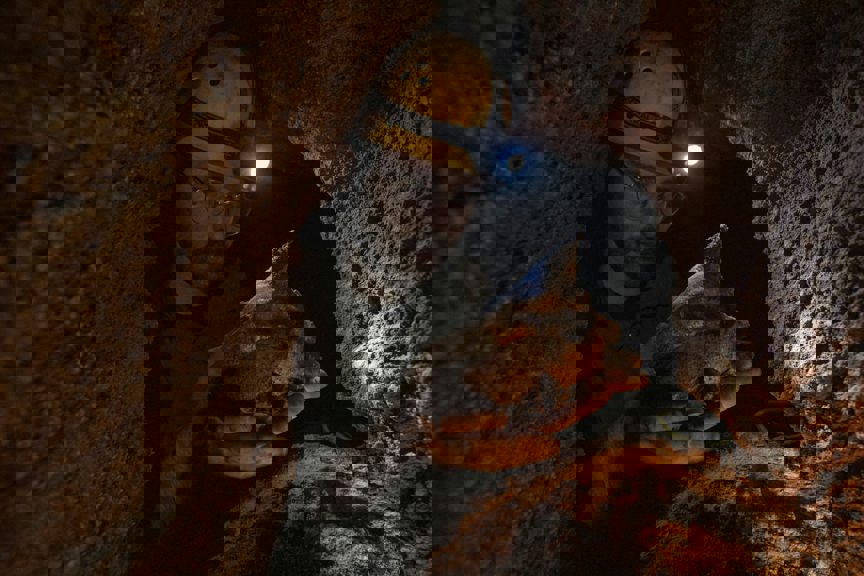 A man in a dark cave wearing glasses and a hard hat with a light on it inspects a large bone