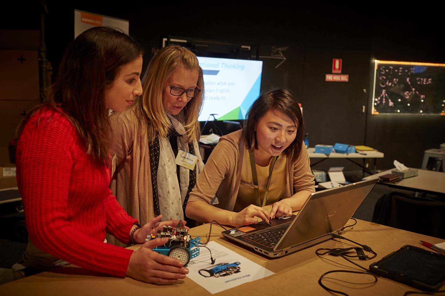 Two female teachers look on as a female museum staff member uses the mouse pad on a laptop. One teacher looks down at a blue car shaped robot with electronics on top.