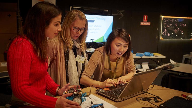 Two female teachers look on as a female museum staff member uses the mouse pad on a laptop. One teacher looks down at a blue car shaped robot with electronics on top.