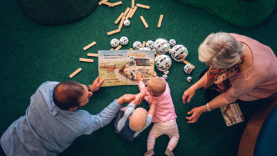 Man and an older lady with two babies laying on the floor looking at a book and objects