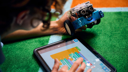 A girl lays on the carpet holding a m-bot robot and programming on an iPad.