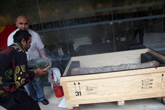 Two men with a bowl of smoking eucalyptus leaves near a box containing part of a carved tree