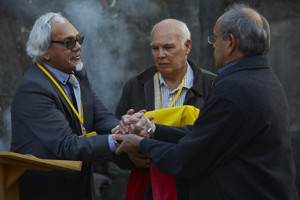 Three men standing in a group. Two clasping hands. On holding a box wrapped in the Aboriginal flag.