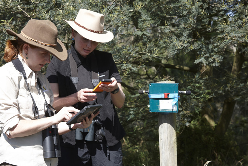 Two women standing in bush land holding mobile devices