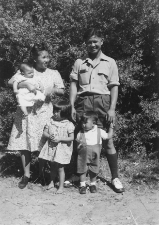 Group of two adults and three children in an outdoor setting, bushes in the background.