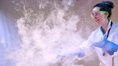 A person wearing a lab coat, safety googles and rubber gloves has lots of white fog around them from pouring liquid nitrogen during a science demonstration.
