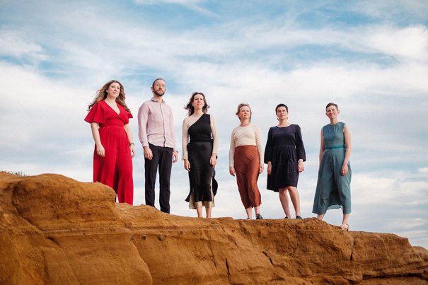 Six members of Alta Collective standing on a sand rock with the cloudy sky behind them.