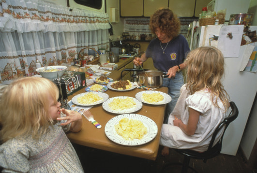 Woman serving food from a saucepan as two small children watch