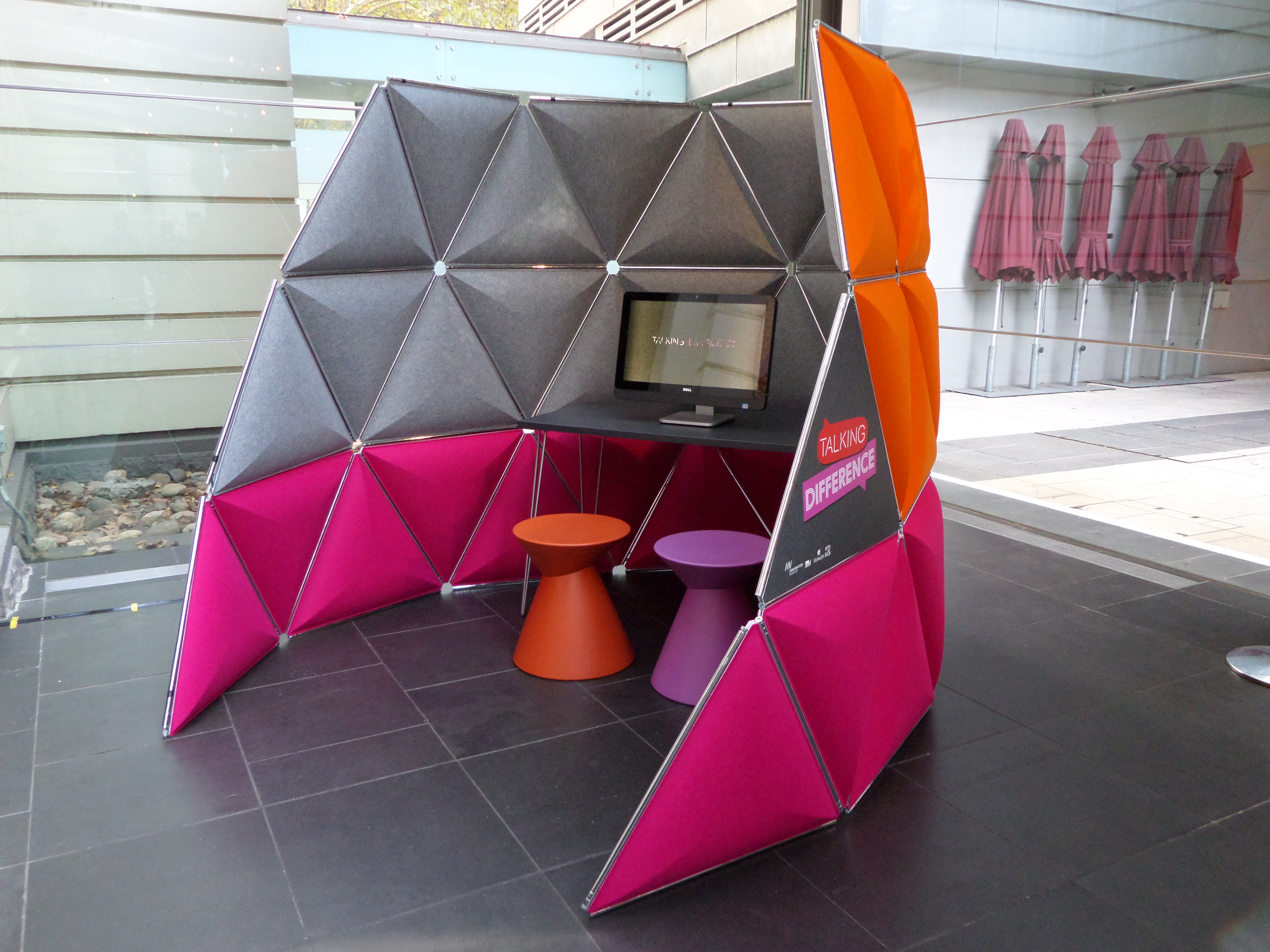 A colourful pod with a computer on a desk with two plastic stools