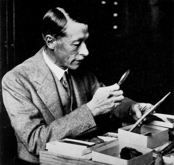 Black and white photograph of a man holding a magnify glass looking at museum specimens