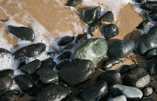Greenish black pebbles on sand surrounded by foam from seawater