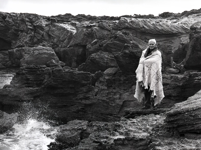 A man stands proudly on a cliff overlooking a wild ocean wrapped in a possum skin cloak.