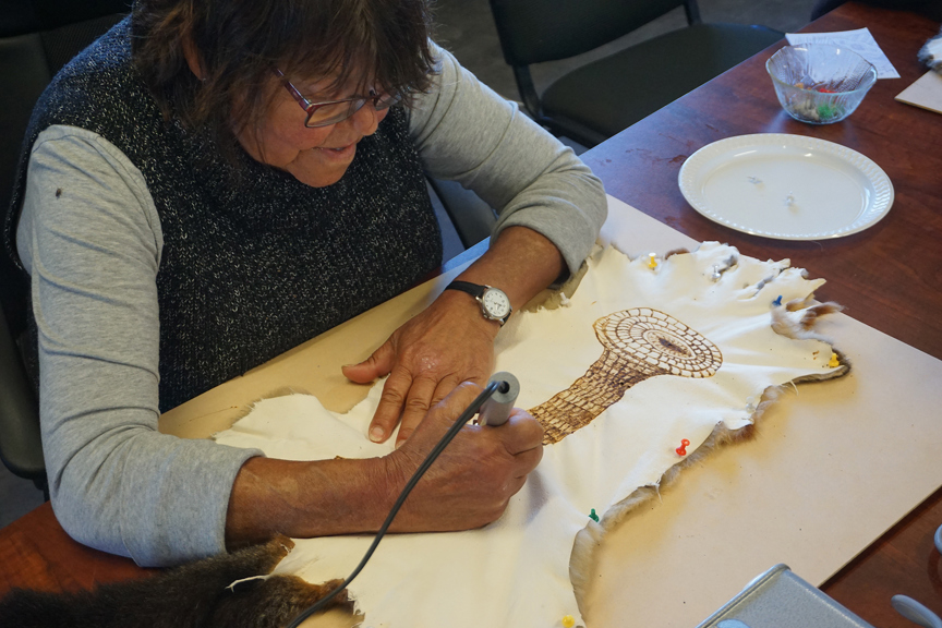A woman sketches an eel basket onto a possum skin with a wire-nib burner.