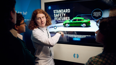 A group of students in the Road to Zero Experience Space standing in front of a touch screen with an image of a green car on the screen 