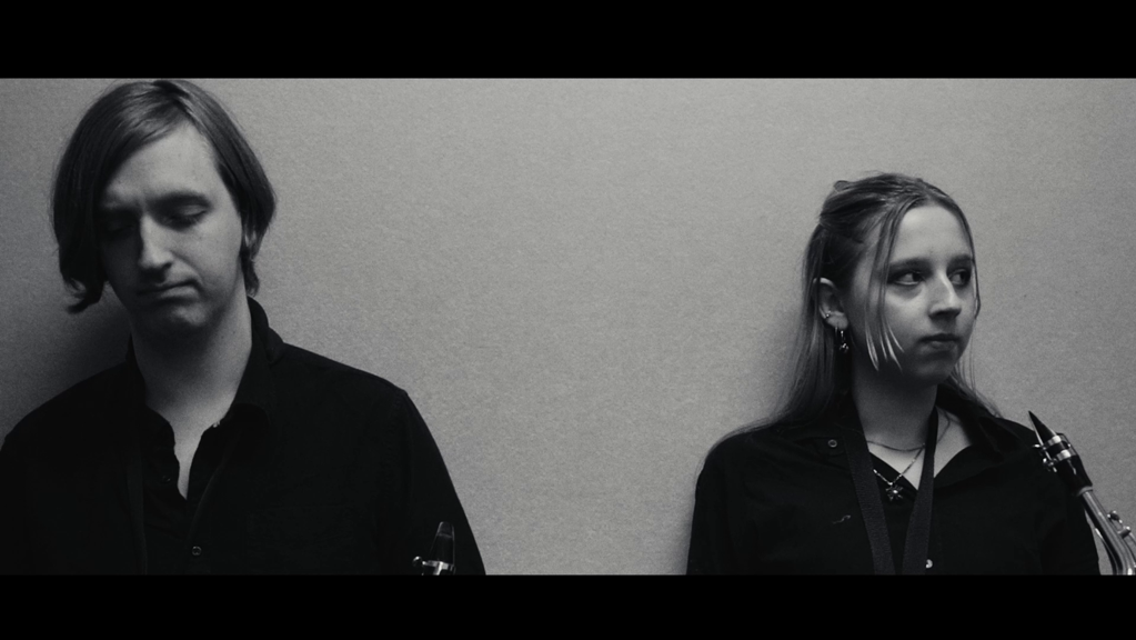 Black-and-white image of two musicians in dark clothing, each holding wind instruments, standing against a plain wall with solemn expressions.