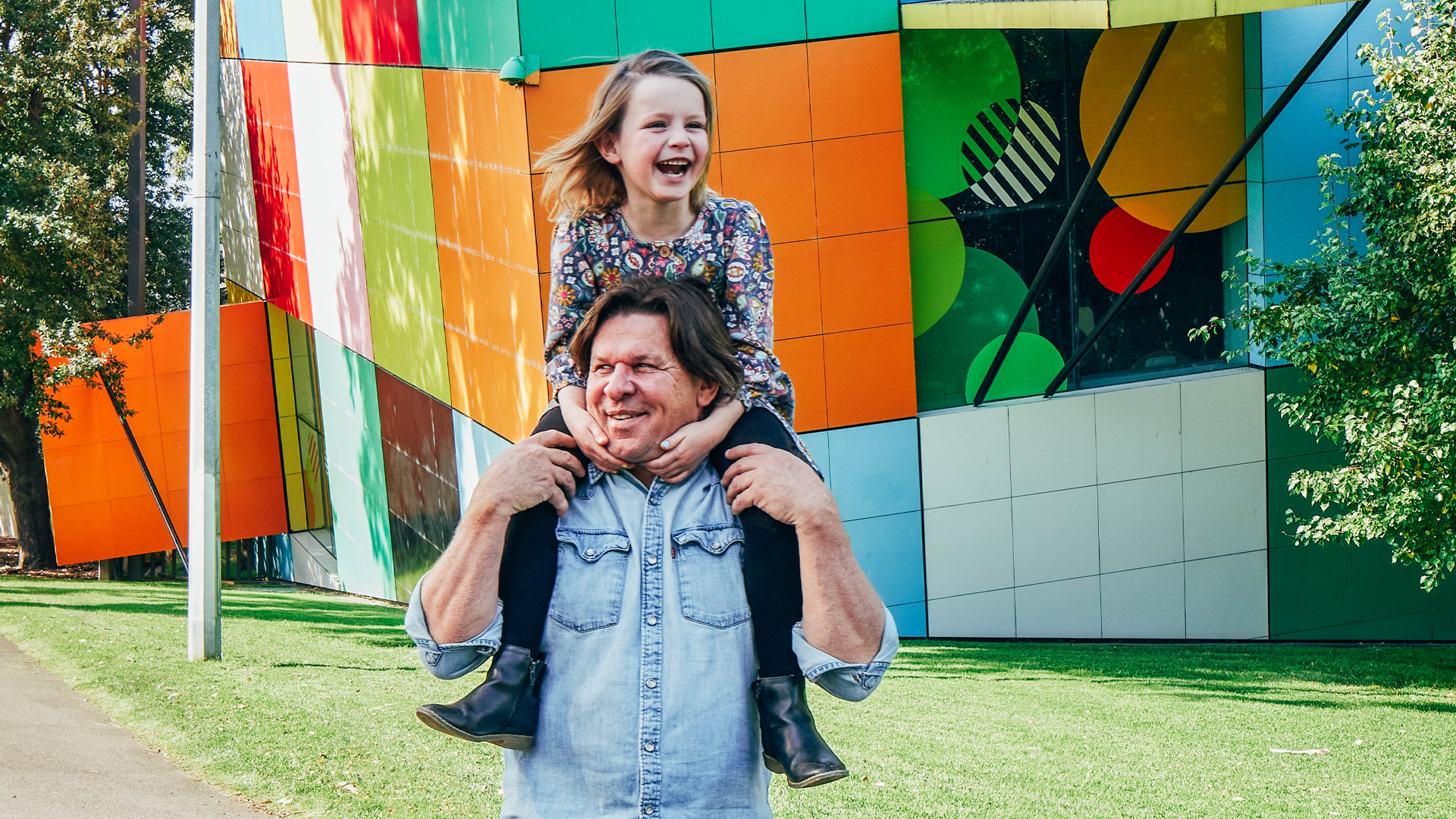 A man with a child on his shoulders walks along a path with the colourful walls to the Children's gallery in the background.