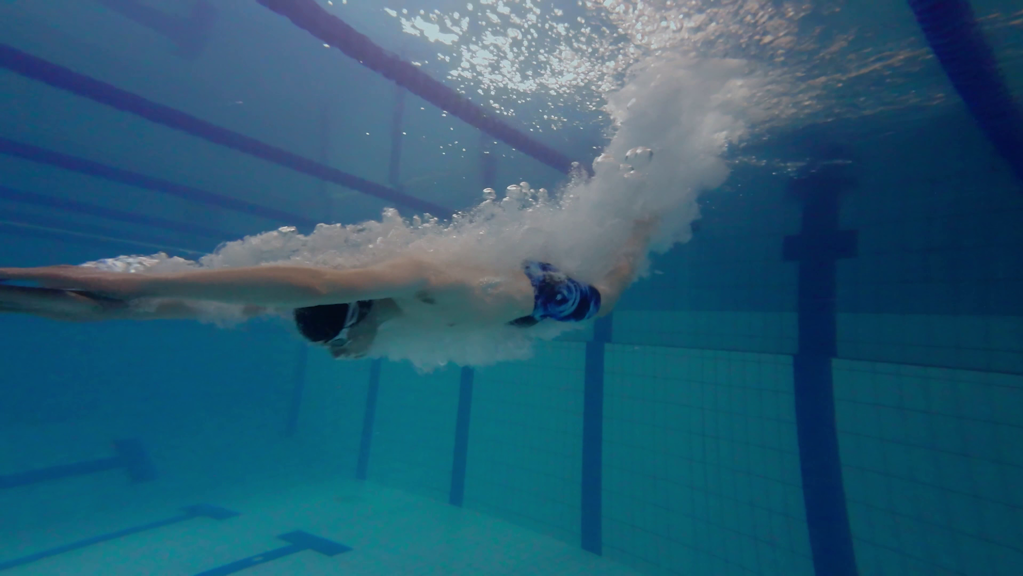 Underwater view of a swimmer wearing goggles and board shorts, diving into a tiled pool.