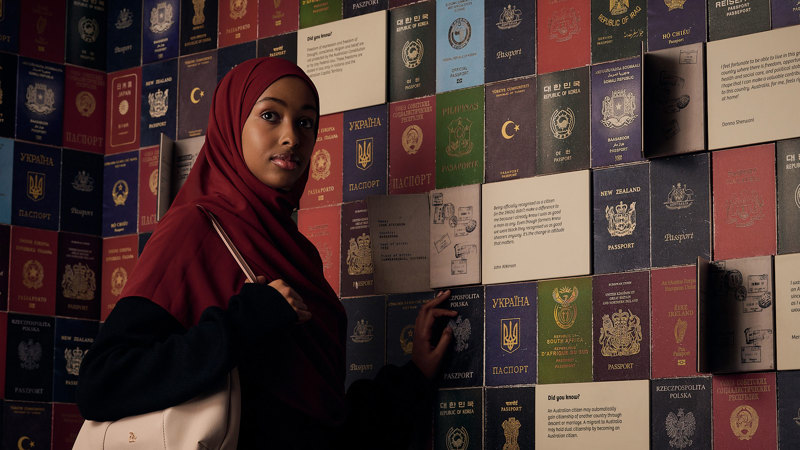 A woman in a hijab stands beside a wall covered in passports from around the world, in Identity: Yours, Mine, Ours exhibition at the Immigration Museum.