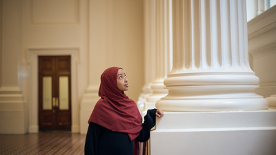 A woman in a hijab leans against a colossal pillar in the Long Room at the Immigration Museum.