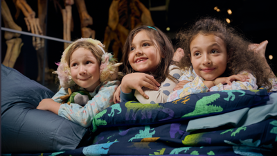 Three children lie on sleeping bags and blankets on the floor of the museum beneath the massive skeleton of a dinosaur.