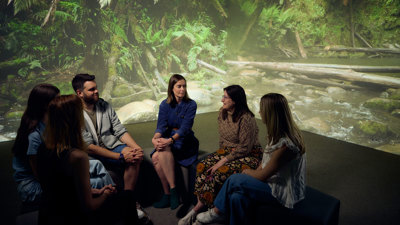 A group of people sitting in a circle having a discussion. A large image of a forest covers the wall in the background.