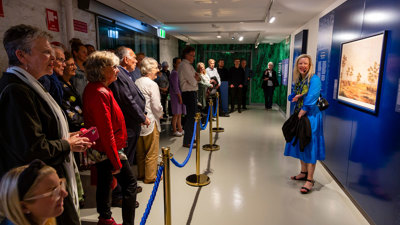A woman in a blue dress stands before displays in the basement of the Royal exhibition Building giving a tour.
