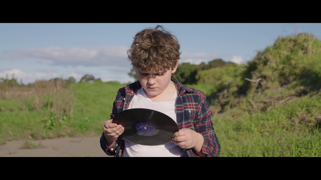 Young boy in a flannelette shirt stands examining a vinyl record, against a backdrop of green hills and blue sky.