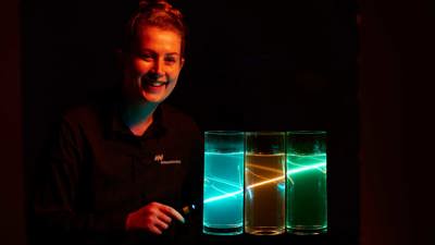 Person with hand-held coloured light source refracting/reflecting in three glass containers filled with liquid durine the Glow Show at Scienceworks.