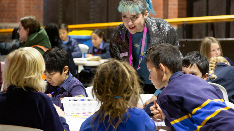 A young adult leans over a table where school students are working. They have pale blue hair and are wearing a pink lanyard that indicates they are a staff member. They look enthusiastic as they talk to the students. 