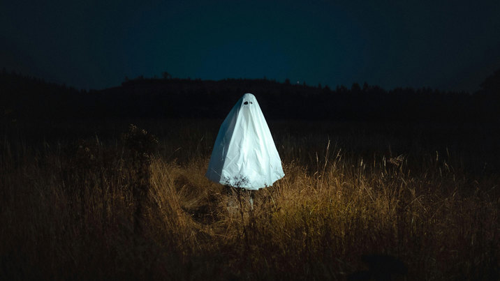 A person wearing a sheet to look like a ghost standing in a field at night.