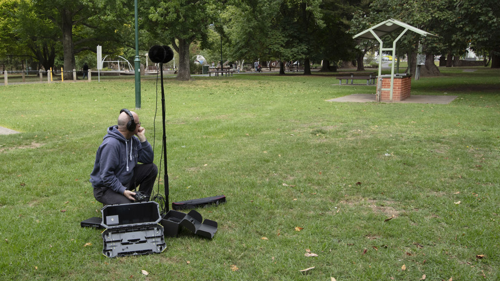 Sound recordist sits on grass besides a large microphone on a stand