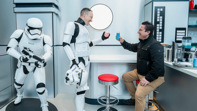 A visitor shares a toast with a Stormtrooper at the Galactic Café, each holding a colourful Star Wars–themed drink, while another Stormtrooper stands nearby.