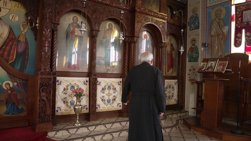 Elderly man in a black priest's robe stands in an ornate church, facing a wall of religious icons and intricate wooden carvings.